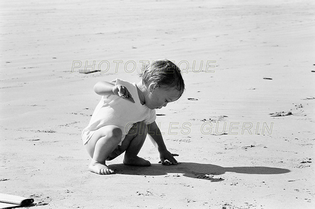 Jeune enfant accroupi pour saisir un coquillage sur une plage de sable en bord de mer - Photo noir et blanc- Presqu-le de Crozon - Bretagne - 1990